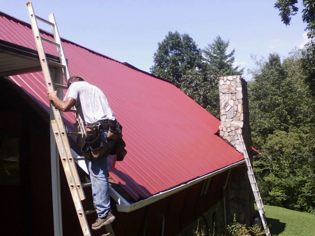 Roofer on a ladder beside a red metal roof and stone chimney — Soffit & Fascia Repair by Roofing Brothers in Elvaston